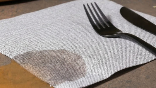A close-up image of an airlaid luncheon napkin shows it cleaning up a small spill on a wooden table, without falling apart like a standard paper napkin would.