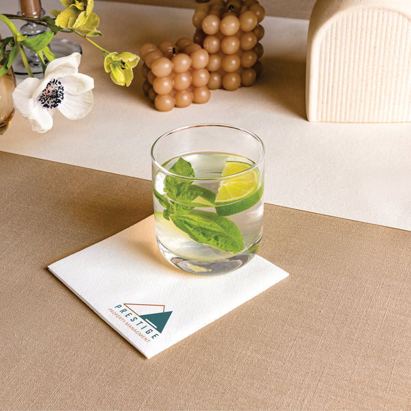 Glass of water with lime and mint leaves on a beige table. The glass sits on a cocktail napkin stamped with a company logo, demonstrating Naptec Paper's customization capabilities.