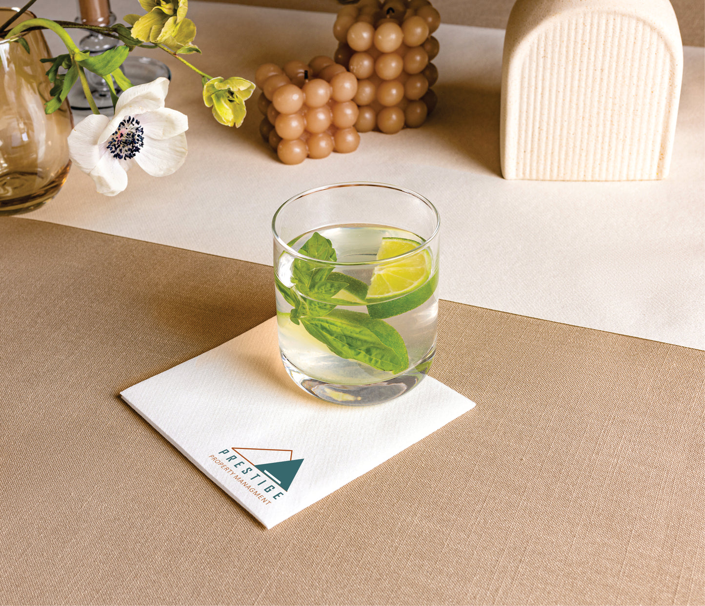 Glass of water with lime and mint leaves on a beige table. The glass sits on a cocktail napkin stamped with a company logo, demonstrating Naptec Paper's customization capabilities.