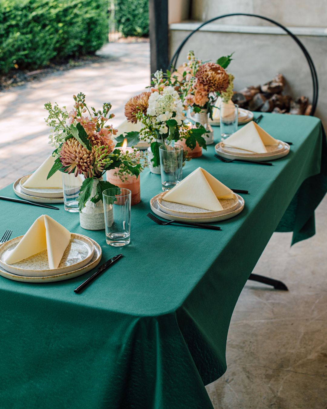 A backyard party setup features Bloomingoods napkins folded in a simple triangle on each place setting. The table is adorned with a Bloomingoods green tablecloth and floral arrangements in white vases.
