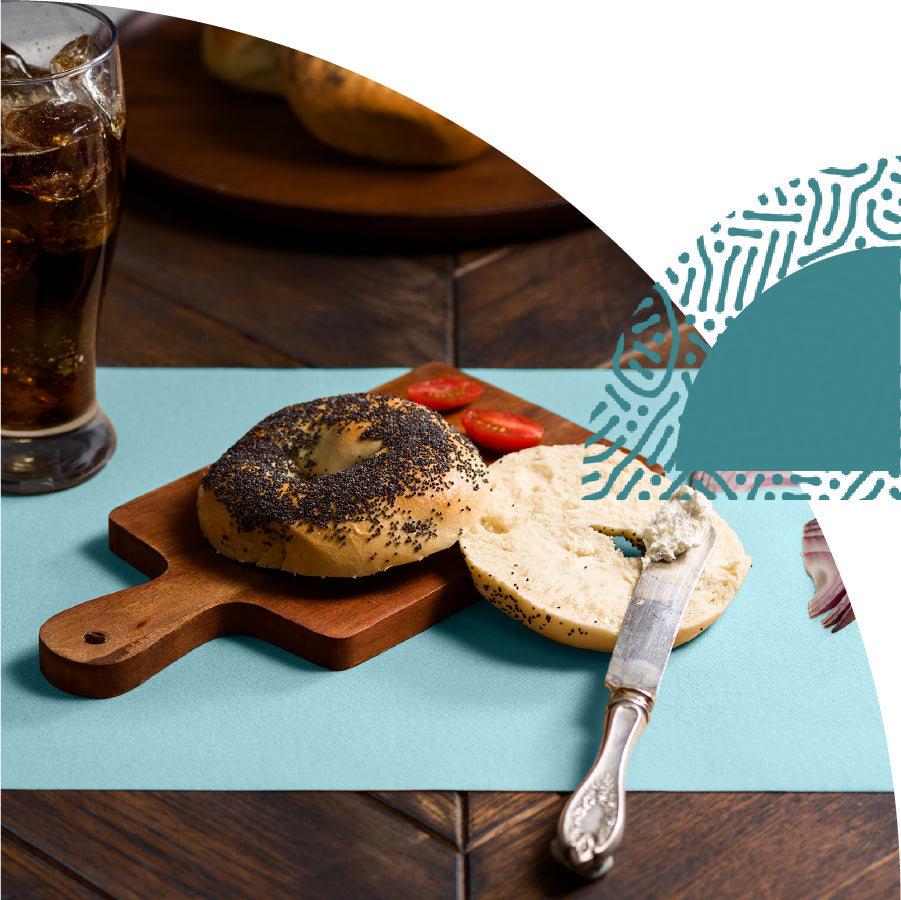 Bagel with cream cheese and a knife on a wooden cutting board with a glass of soda in the background.