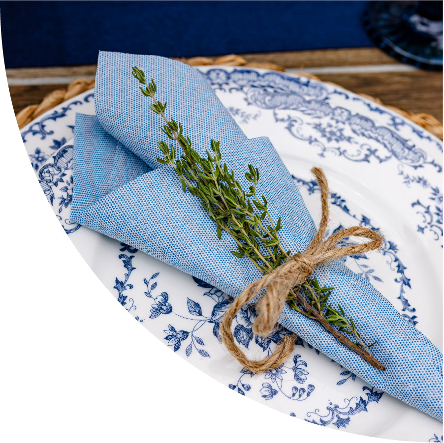 A dusty blue disposable napkin from Naperee is tied together with twine. The bundle includes a sprig of thyme. The bundle is set on a decorative plate.