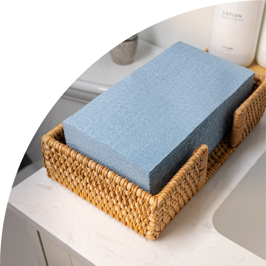 A stack of dusty blue colored hand towels in a woven basket. The basket is sitting on a bathroom sink. The single use high quality guest towels are from Naptec.