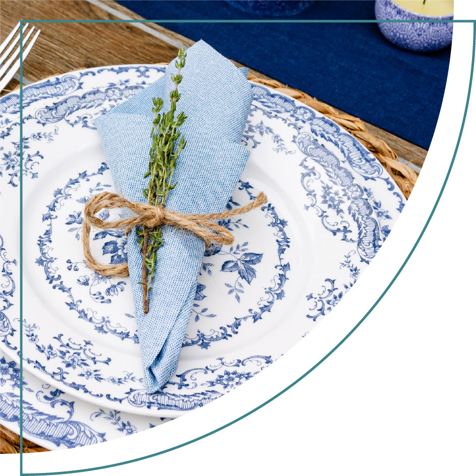 A dusty blue napkin sits on a blue and white plate. The Naperee napkin is folded into a cone shape and tied into a bundle with twine. There's a sprig of thyme in the bundle.