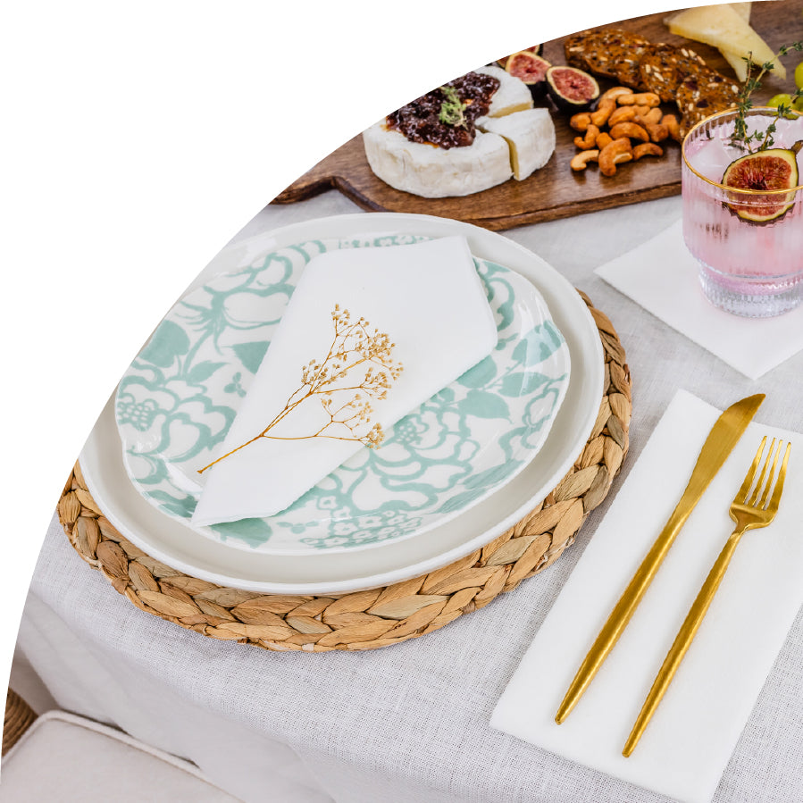 Table setting with floral-patterned plates, gold cutlery, and a small cheese board with brie, figs, and nuts. The white napkin on the dishes is from Lintext.