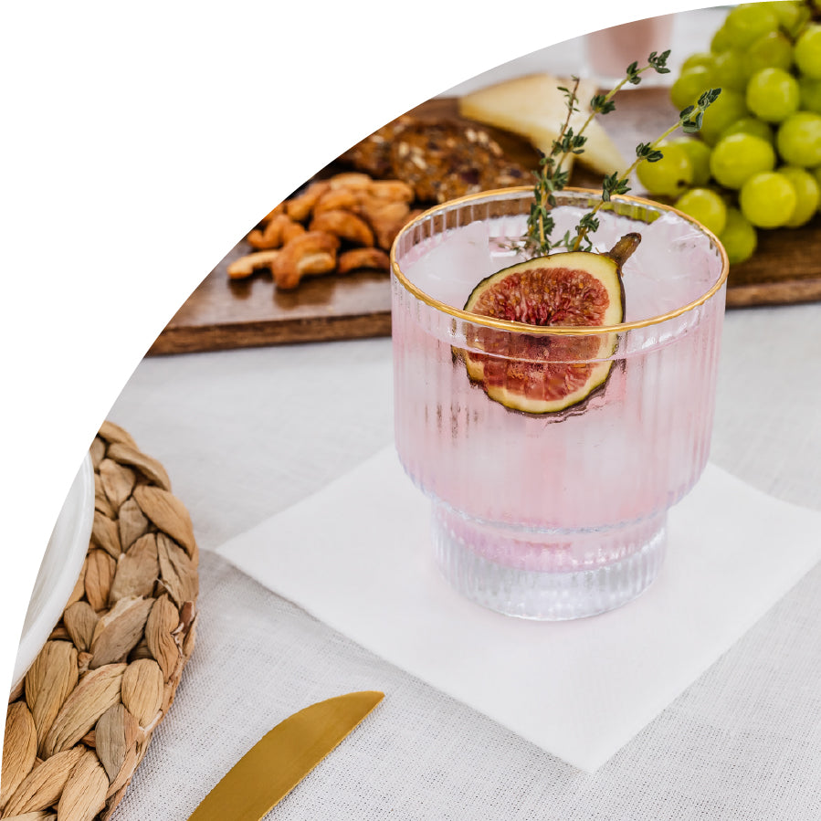 Pink cocktail with figs in a glass, surrounded by grapes and nuts on a white disposable cocktail napkin from Lintext.