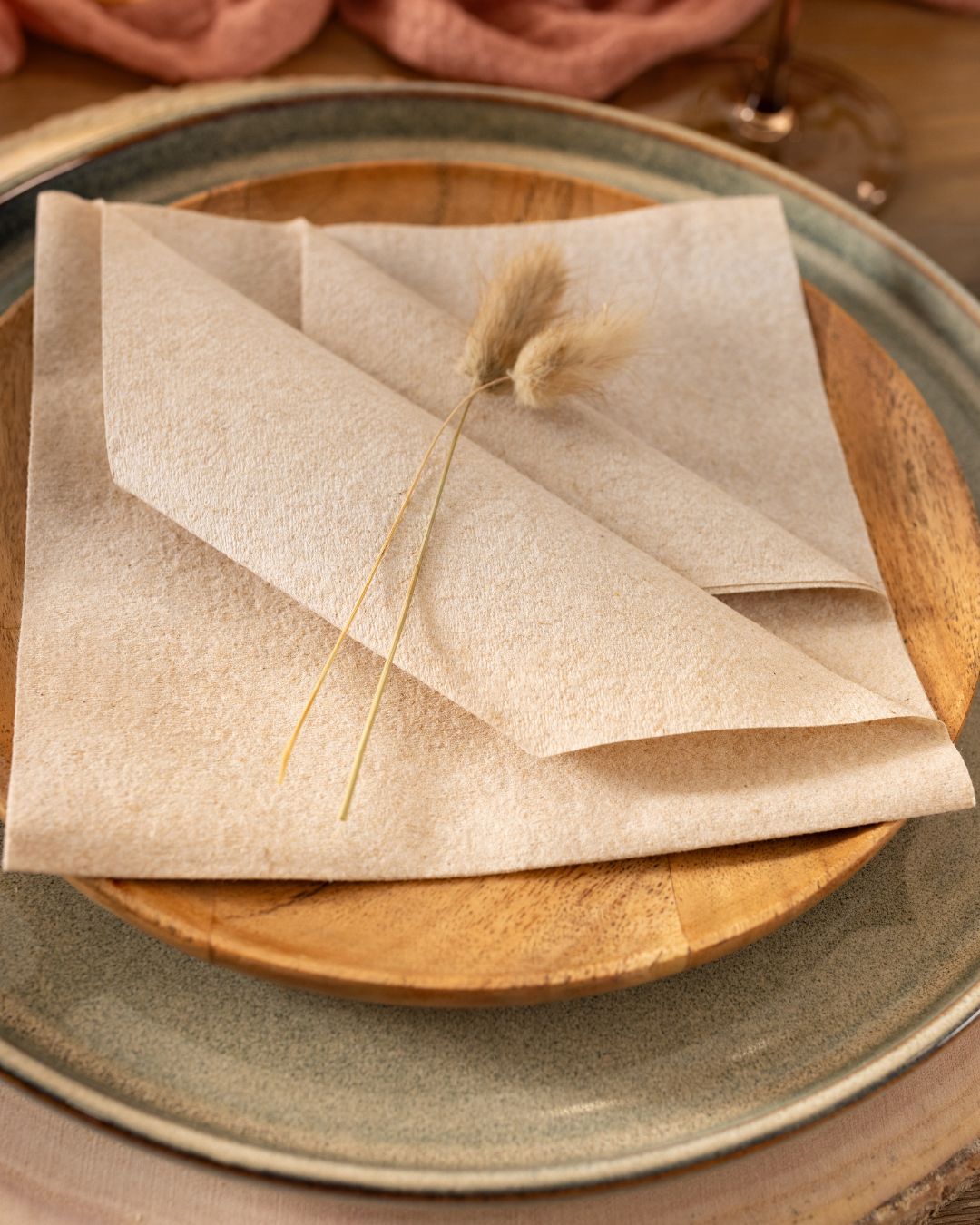 A Servietto tan disposable napkin is folded twice in a basic fold. It lays on a wooden place setting. Two faux wheat chaffs lay on top of the naptec.