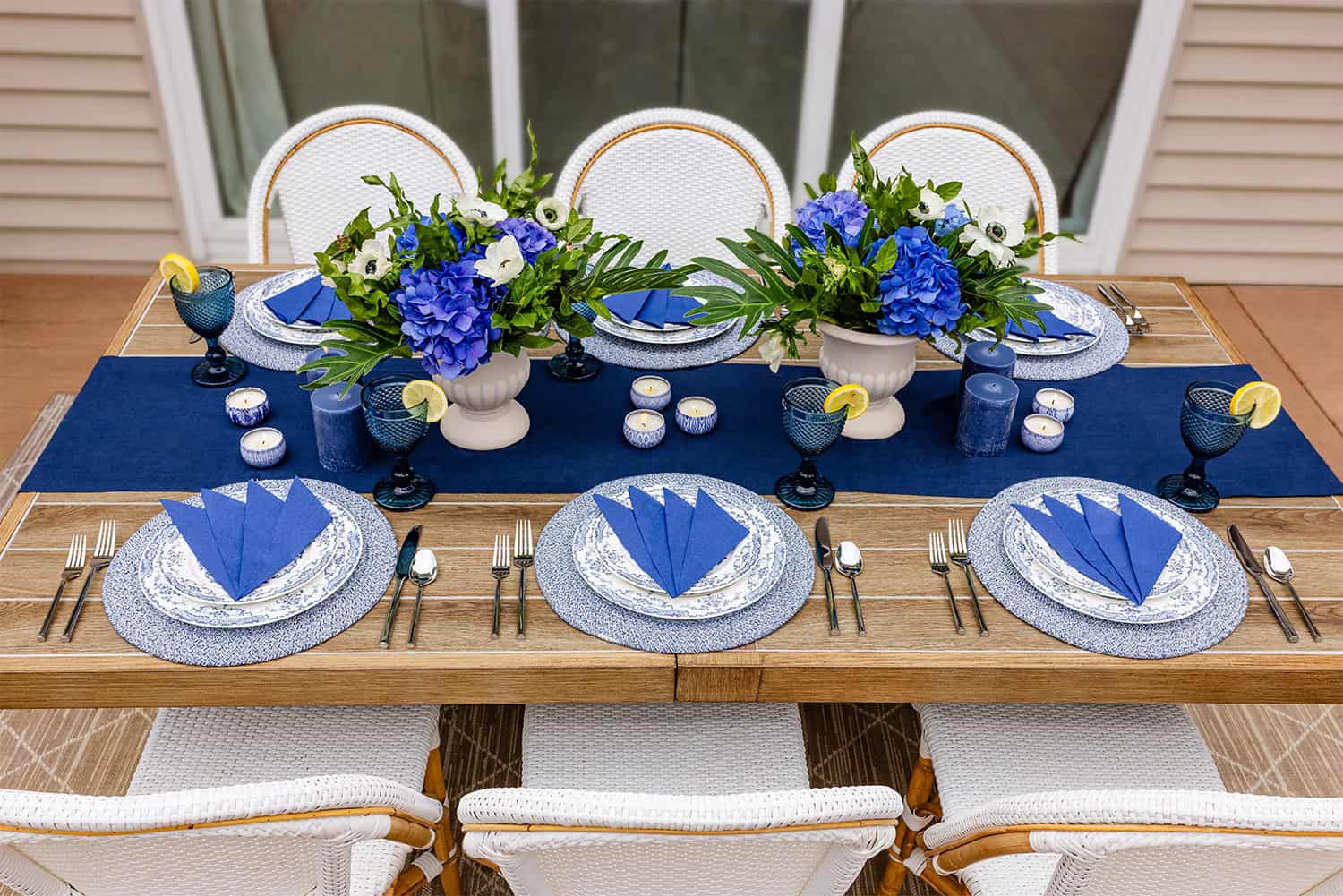 An outdoor table setting features cornflower blue paper apkins from Naptec Paper and a deep blue table runner from Servietto. There are six place settings, with purple, blue, white, and green floral arrangements in the middle. 