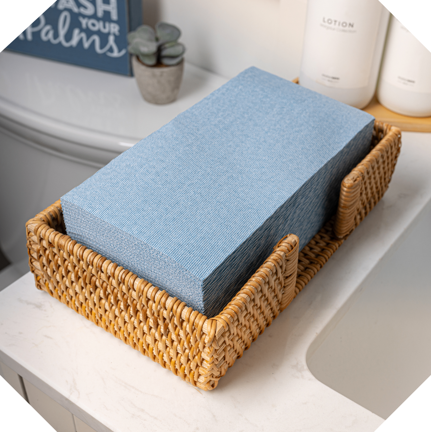 A stack of Lintext disposable guest towels is in a woven guest towel basket. The basket is on a sink in a hotel.