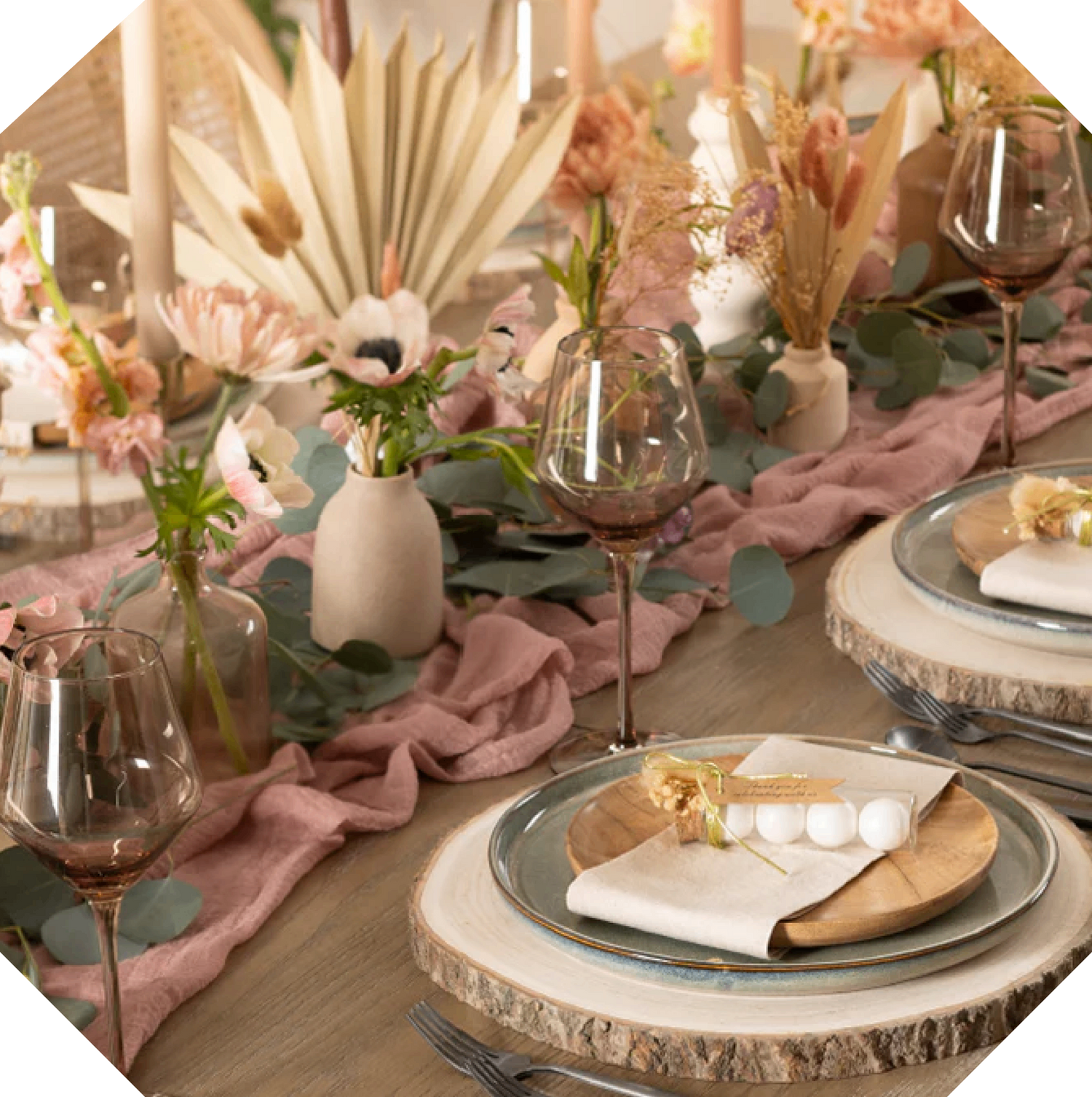 A Naperee single use dinner napkin is wrapped around a place setting. The place setting is layered with wood accents. Centerpiece with pink table runner and eucalyptus leaves runs down the center of the table.